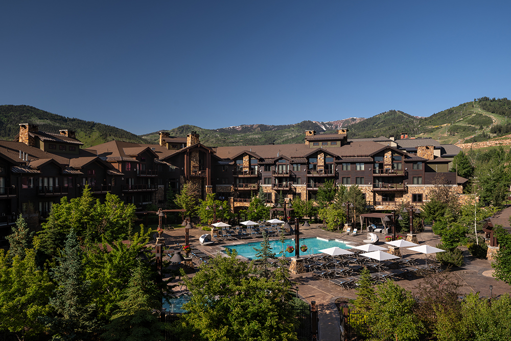 A luxury mountain resort nestled among forested hills, featuring multiple chalet-style buildings, two expansive outdoor pools, and plenty of shaded lounge chairs under white umbrellas. The backdrop includes lush green slopes and a clear blue sky.