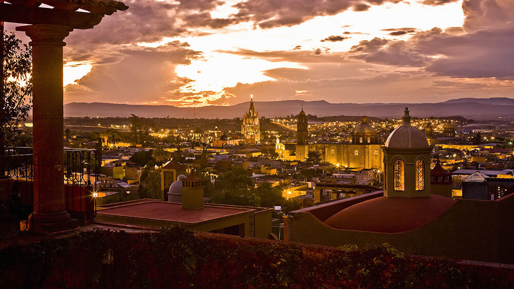 A breathtaking sunset view over San Miguel de Allende, Mexico, featuring the iconic Parroquia de San Miguel Arcángel illuminated in the distance. The foreground includes a charming terrace with colonial architecture and a domed rooftop. The sky glows in warm hues of purple, orange, and gold, casting a romantic ambiance over the historic town and surrounding landscape.