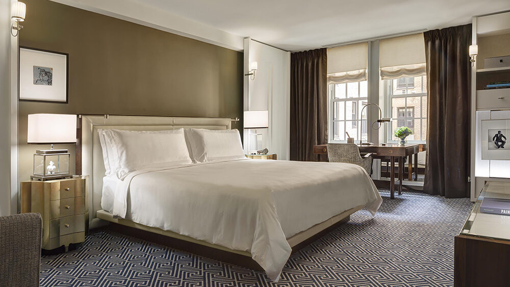 Elegant hotel room featuring a large king-size bed with crisp white linens, flanked by modern bedside tables and soft lighting. A writing desk sits near tall windows draped with dark curtains, allowing natural light to illuminate the sophisticated neutral-toned interior.