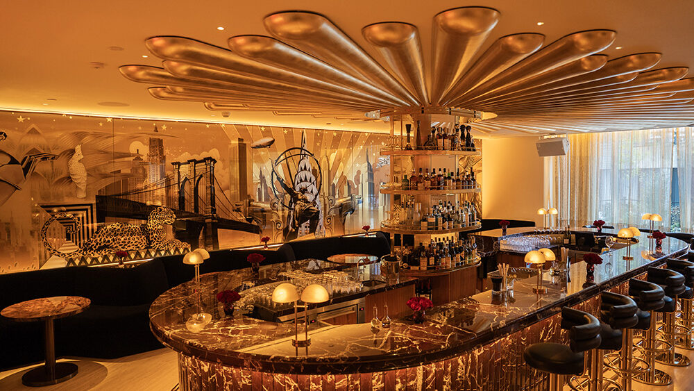 A glamorous, warmly lit bar interior featuring a dramatic gold ceiling installation shaped like a giant flower radiating from the central bar. The bar itself is finished with rich, dark marble and surrounded by high black leather stools with gold bases. Small lamps and red rose arrangements add elegance to the countertop. In the background, a mural-style wall art displays iconic city elements, including bridges, skyscrapers, and a leopard, giving the space an eclectic and sophisticated atmosphere. Large windows on the right allow soft daylight to filter through sheer curtains.