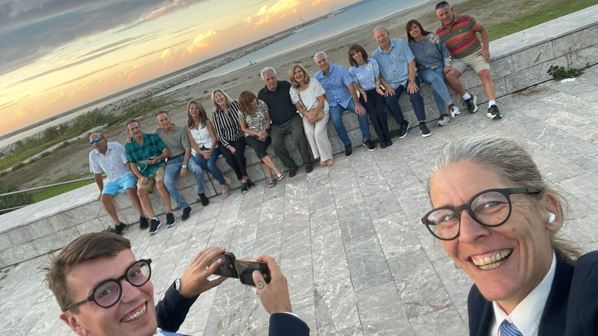 Inspirato team members taking a joyful selfie in front of a large family group during sunset on the Sicilian coast, capturing a moment from a bespoke reunion experience.