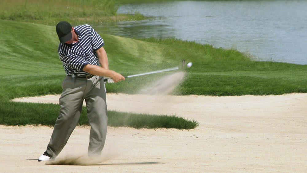 A golfer in a striped shirt and khaki pants hitting a shot from a sand bunker on a golf course. Sand sprays into the air as the club makes contact with the ball, which is seen in mid-flight. The background features manicured green grass and a calm water hazard surrounded by natural foliage.