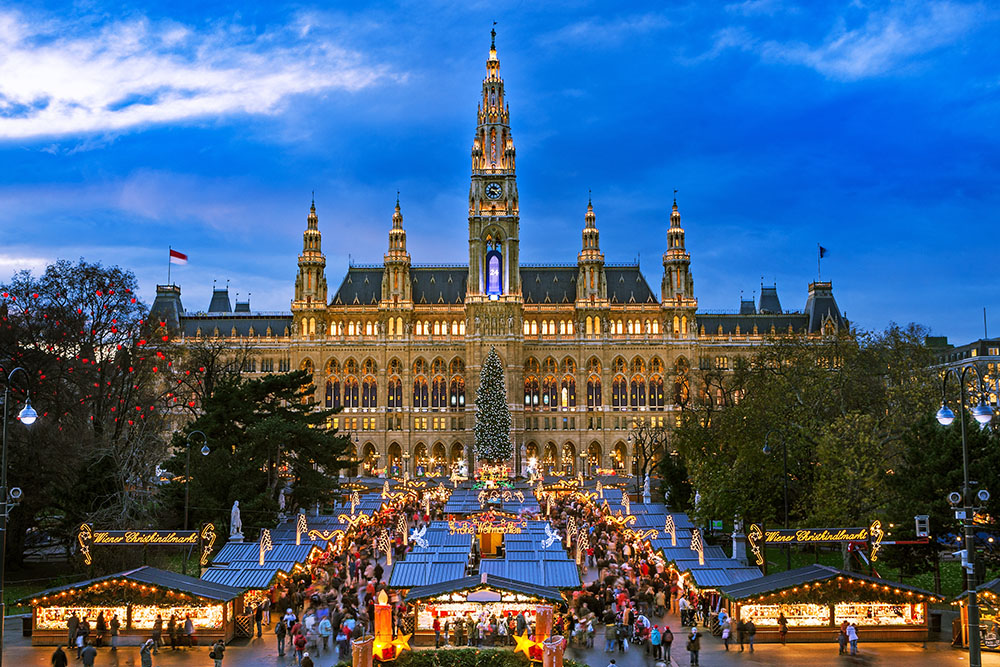 A vibrant evening scene of the Vienna Christmas Market (Wiener Christkindlmarkt) in front of the illuminated Vienna City Hall (Rathaus). The market is bustling with visitors exploring rows of festive wooden stalls, a tall Christmas tree stands in the center, and holiday lights and decorations create a warm, magical glow against the twilight sky.