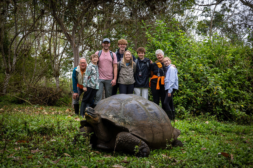 A multi-generational Inspirato member family posing with a Galápagos giant tortoise in the lush highlands of the Galápagos Islands, Ecuador. A memorable wildlife encounter during a Silver Origin luxury expedition cruise, ideal for family travel and eco-luxury exploration in South America.