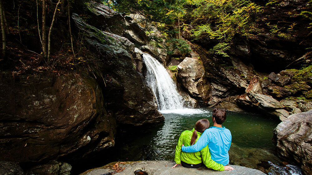Couple sitting beside Bingham Falls near Stowe, Vermont surrounded by mossy rocks and forest with cascading waterfall flowing into a clear pool