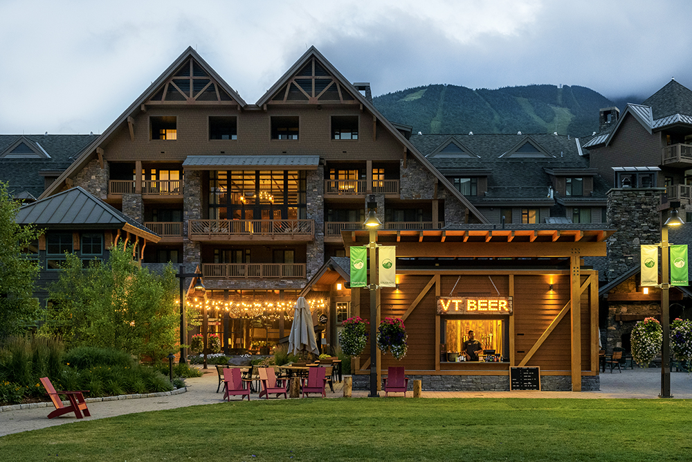 Evening view of The Lodge at Spruce Peak in Stowe, Vermont with illuminated VT Beer kiosk, string lights, and mountain backdrop
