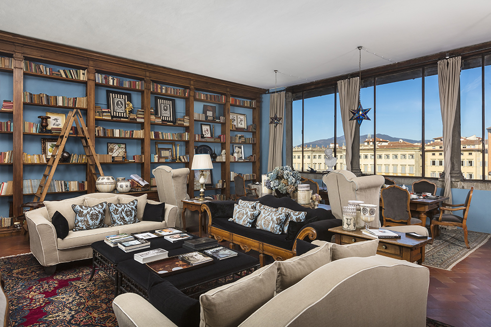 Bright and airy Florence villa living room with designer furniture, classic fireplace, neutral-toned armchairs, and open double French doors leading to a lush private garden.