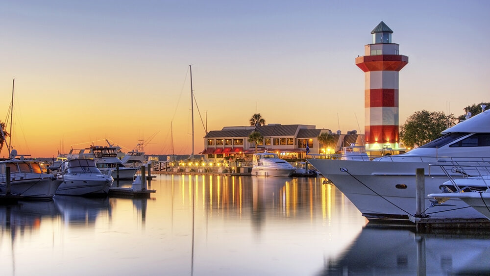 Sunset at the harbor in Hilton Head, South Carolina featuring yachts, waterfront dining, and the iconic red-and-white striped lighthouse, capturing a relaxing spring break atmosphere.