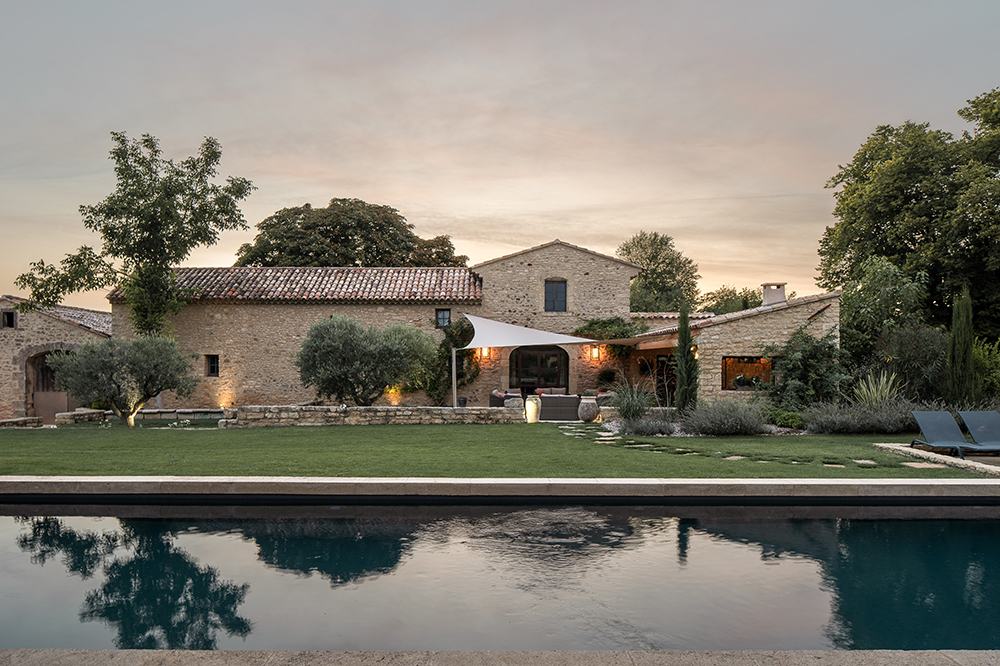 Stone farmhouse with terracotta roof and shaded outdoor seating area beside a tranquil swimming pool in Provence at sunset.