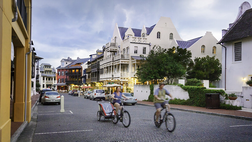 Charming street scene in Rosemary Beach, Florida with pastel-colored buildings and families riding bikes along cobblestone roads, ideal for a walkable luxury spring break destination.