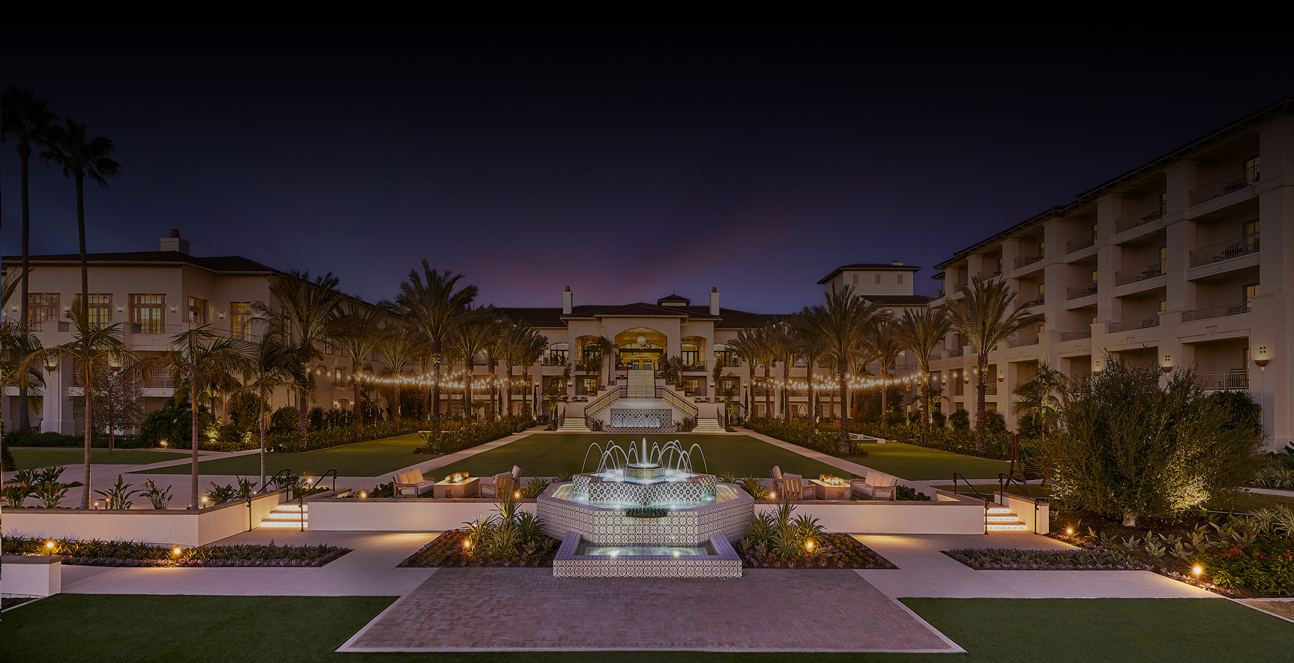 Grand Mediterranean-style resort courtyard at sunset, featuring a glowing tiered fountain, manicured lawns, palm-lined walkways, and warm terrace lighting beneath a vibrant twilight sky.