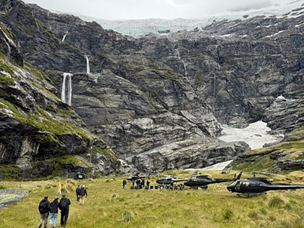 Inspirato members walking toward helicopters in a remote alpine valley at Earnslaw Burn, surrounded by waterfalls, steep rock faces, and a glacier under cloudy skies.