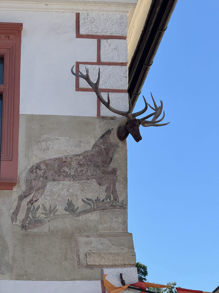 Mounted stag antlers extend from the corner of a historic building façade, positioned above a faded mural of a deer painted on the stucco wall, set against a clear blue sky.