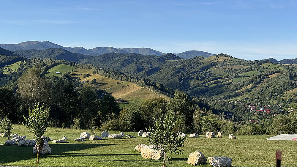 Rolling green hills and forested mountains stretch across the Transylvanian countryside under a clear blue sky, with scattered stone accents and young trees dotting a manicured lawn in the foreground and a small village nestled in the valley below.