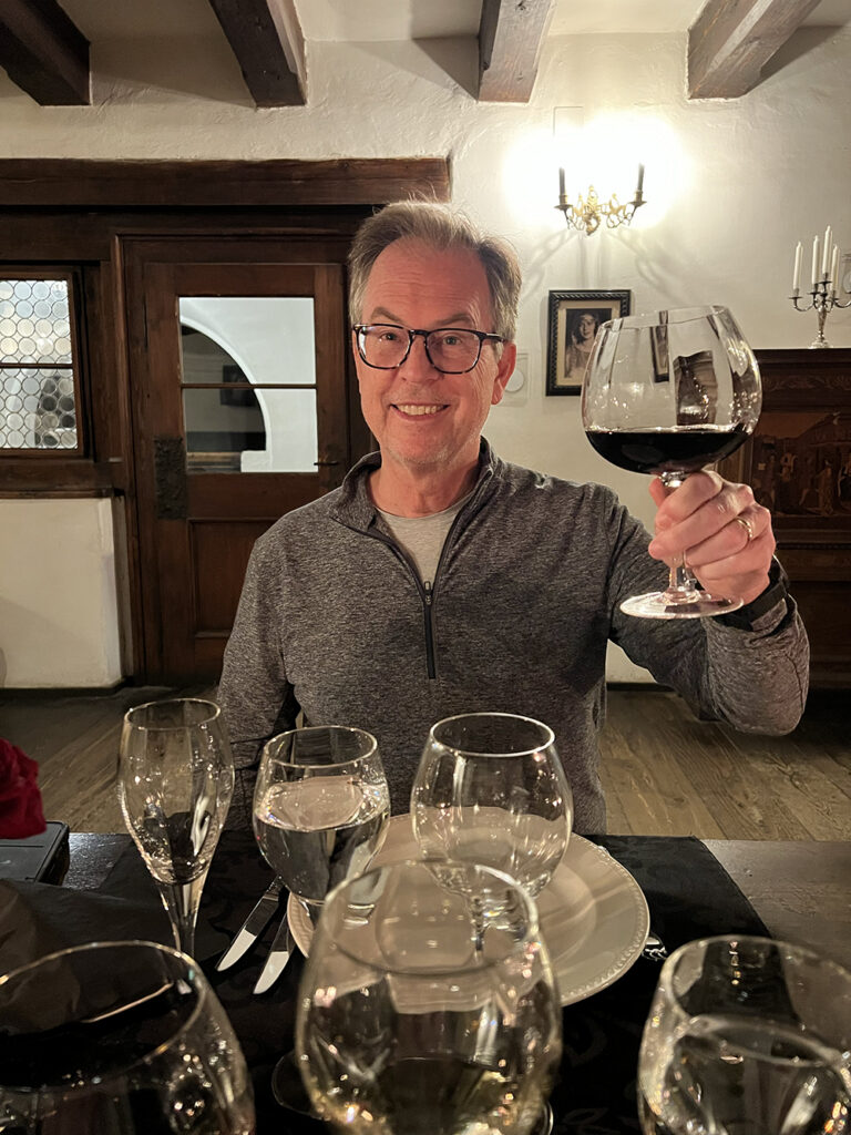 A man seated at an elegant dining table inside a rustic, candlelit restaurant raises a large glass of red wine in a toast, surrounded by polished glassware, wooden beams, and warm ambient lighting.