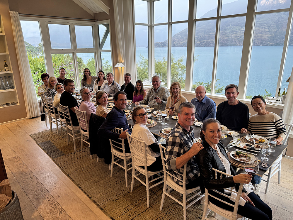 Inspirato members gathered around a long dining table enjoying a group dinner with panoramic lake and mountain views in Queenstown, New Zealand.