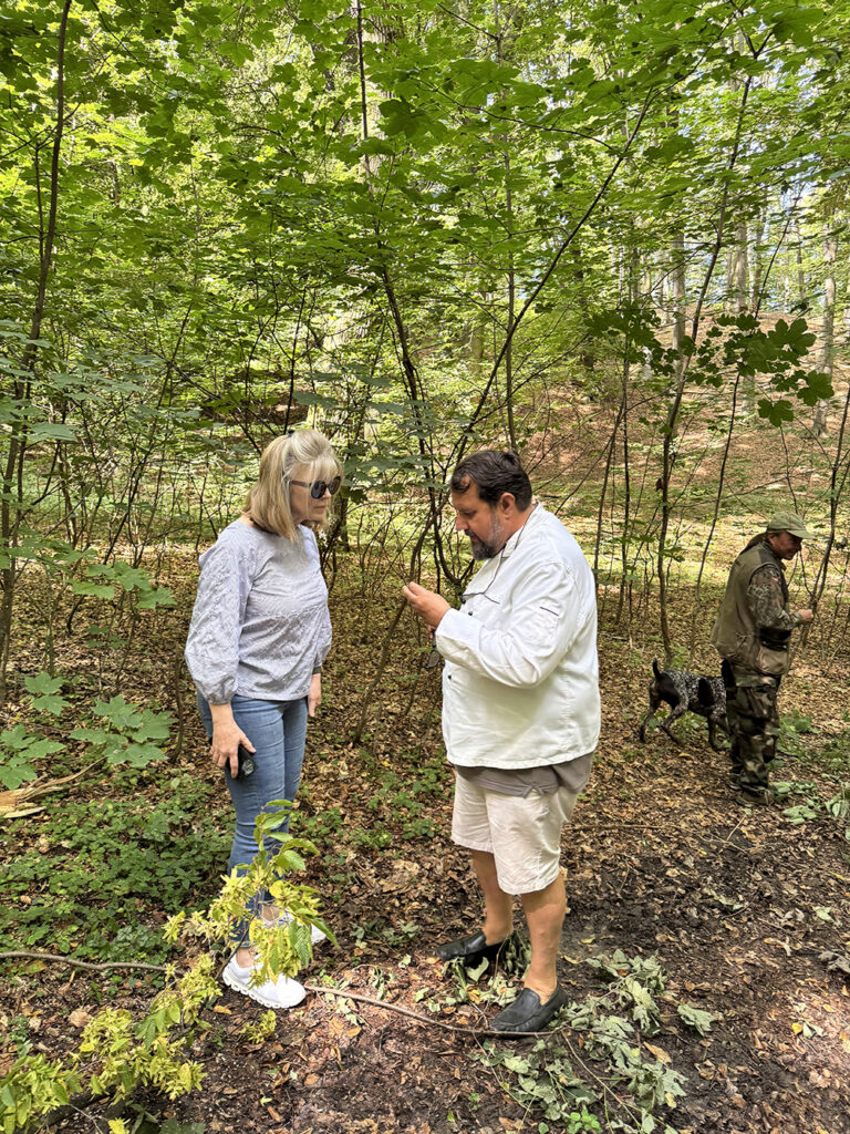 In a lush forest setting, Shelly speaks with a local guide who holds a small object for closer inspection, while another man and a dog move through the trees in the background, suggesting a hands-on countryside experience.