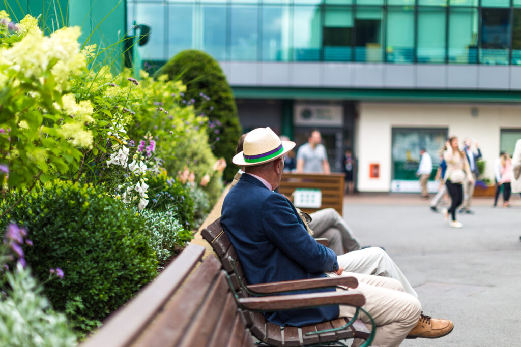 an elderly man wearing a hat sits on a park bench outside centre court
