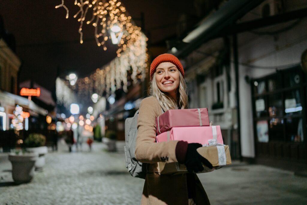 A young woman walking through the city at Christmastime, carrying a stack of wrapped gifts and smiling